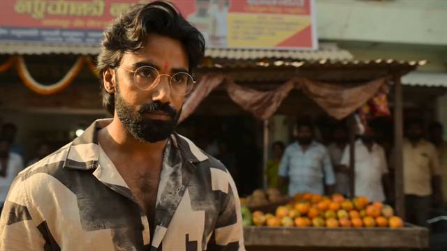 Close-up of a bearded man in round glasses in front of a small-town market — painted shop signage, fruit stalls and onlookers behind him.